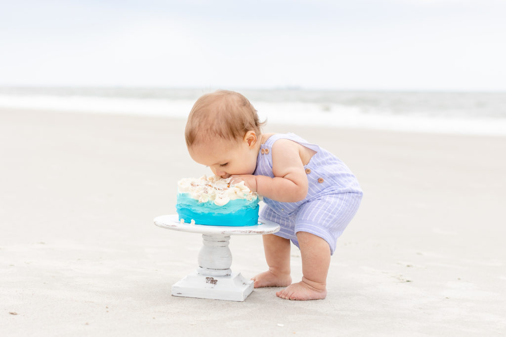 Baby boy wearing blue eating a blue and white cake on the beach.