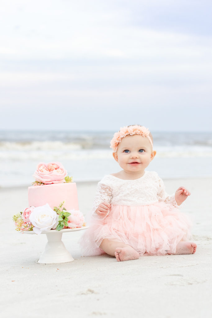 Baby girl wearing a pink dress and a pink flower headband is sitting next to a pink cake with flowers on the beach.