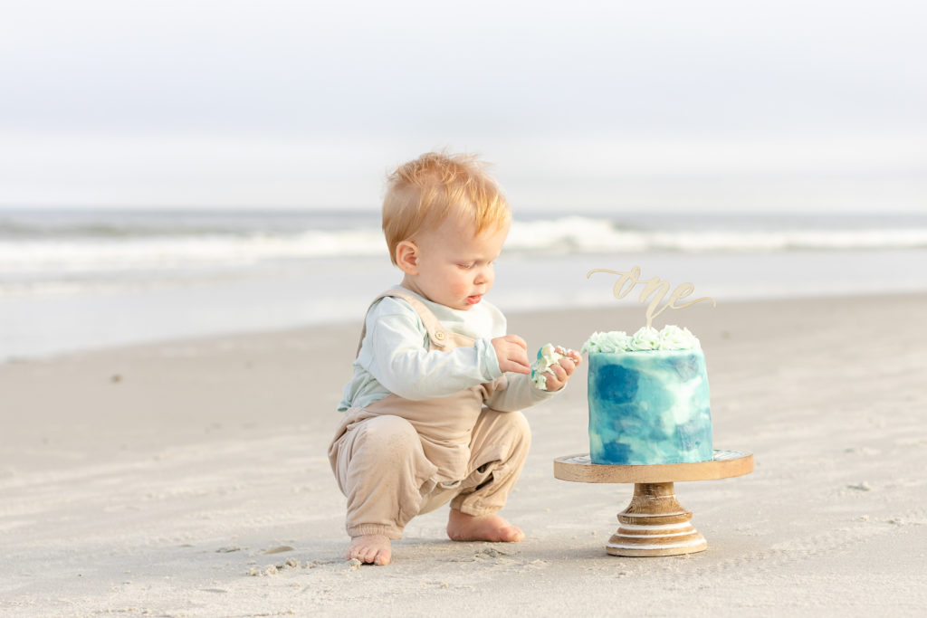 Baby boy playing with blue cake that has been placed next to him. The cake has a decoration on the top that says one. The baby and the cake are on the beach.