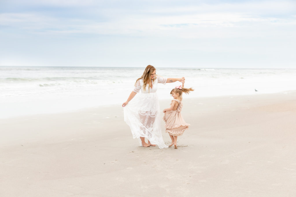 A family is on the beach. The mom is wearing a white flowy dress and is twirling her daughter who is wearing a pink dress. They are standing in front of the ocean.