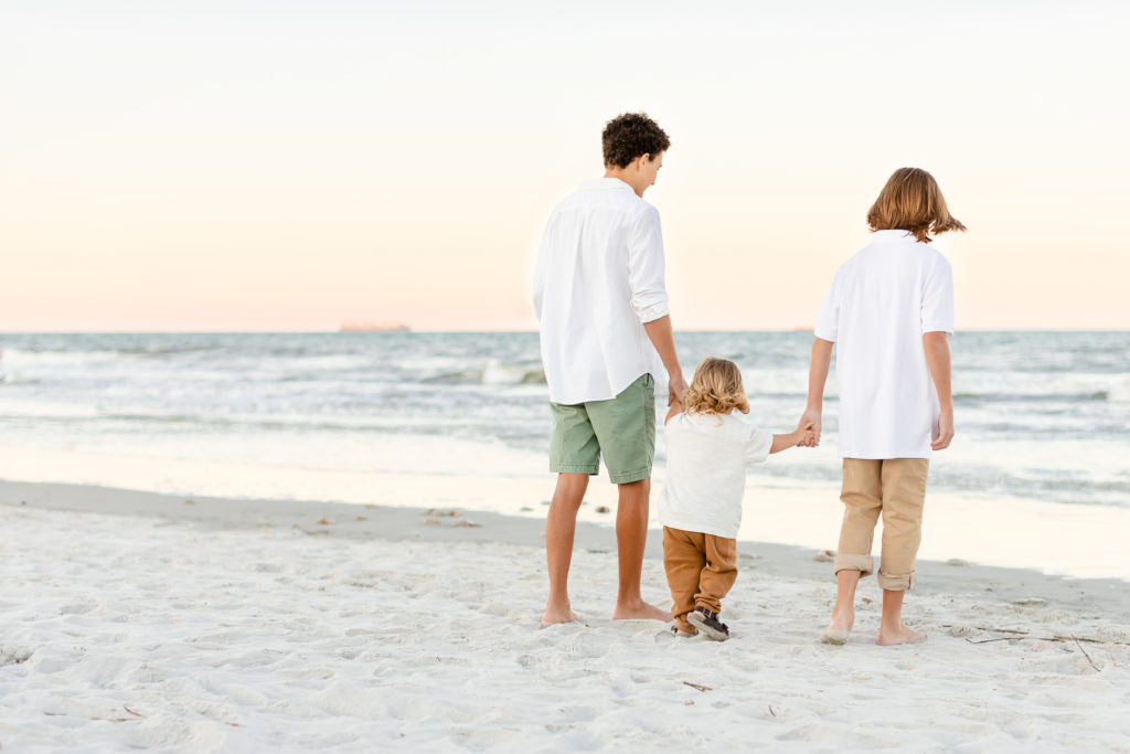 Three brothers are standing on the beach looking into the ocean. They are all holding hands wearing white shirts.