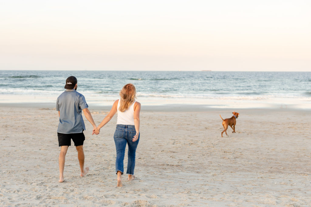 There is a family on the beach. The husband and wife are holding hands walking towards the water. Their brown dog is running towards the ocean.